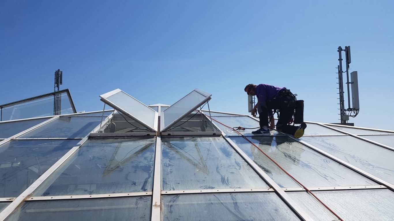 Un homme nettoie le toit en verre d'une serre sous un ciel ensoleillé.