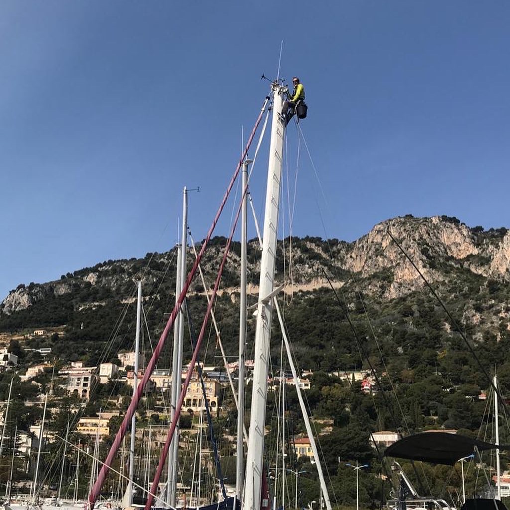 Un homme en vêtement de sécurité grimpe à un mât dans un port avec des montagnes en arrière-plan.